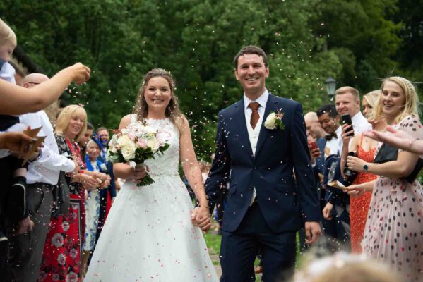 A professional wedding photograph of a husband and wife walking towards the camera while their friends and family throw confetti over them.