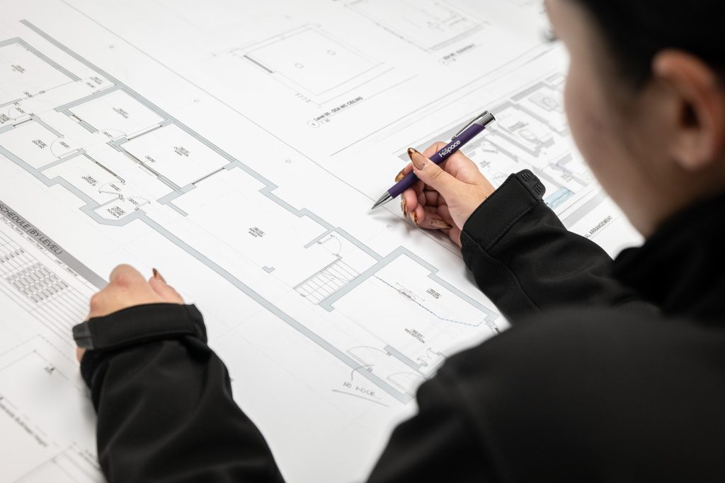 A professional studies detailed blueprints and makes notes with a pen in a technical office setting.