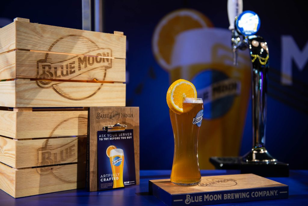 Pint of Blue Moon beer with an orange slice, displayed beside branded wooden crates and a bar tap.