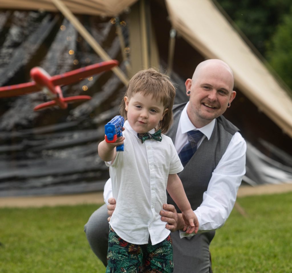 A young boy in a white shirt and bow tie points a blue toy gun with a red toy plane mid-air, while his smiling dad crouches behind him outside a tipi.