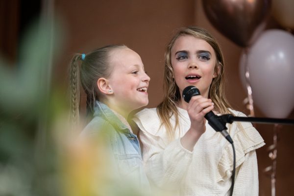 Two young girls singing into a microphone at a party or talent show, with balloons and decorations in the background.