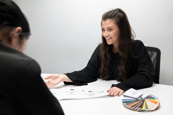 A smiling consultant presents design options to a client across a table, with colour swatches visible in the foreground.