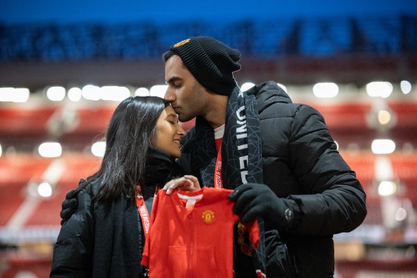 A couple embrace at a football stadium, holding up a baby onesie as they announce their pregnancy.