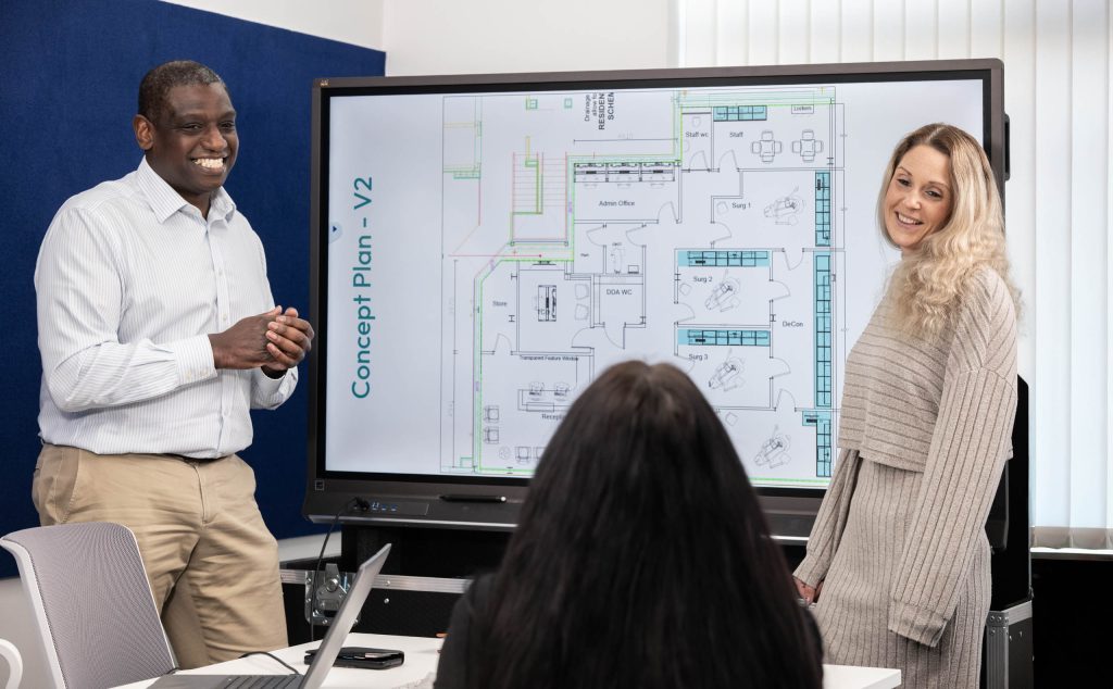 Two presenters lead a planning session with a large digital screen showing a dental clinic concept plan.