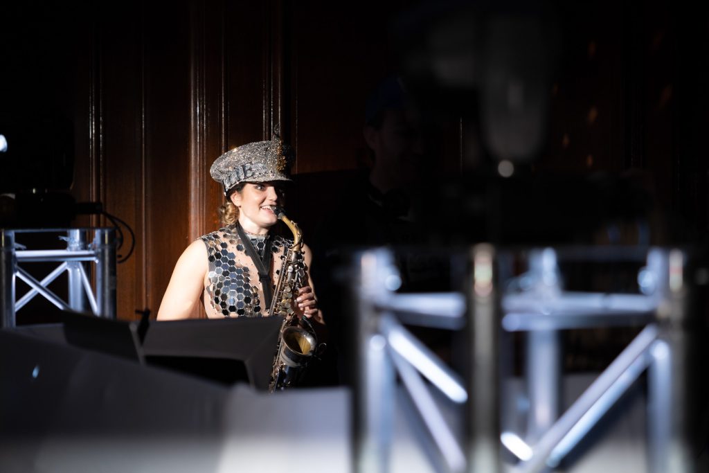 Saxophonist in a sequinned outfit and glittery hat, smiling while lit by a spotlight during a performance.