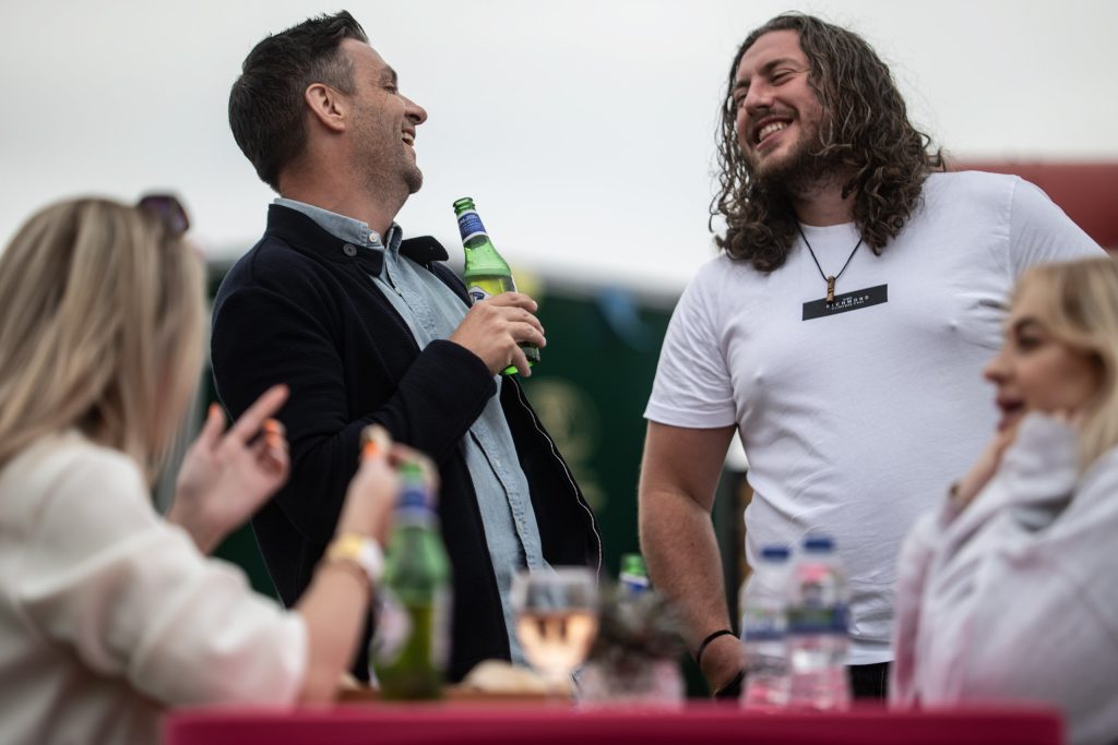 Two men laughing while holding drinks, surrounded by friends at a table in a relaxed outdoor setting.