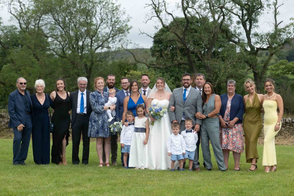 Large wedding party including bride, groom, children and relatives gathered on a lawn with countryside in the background.