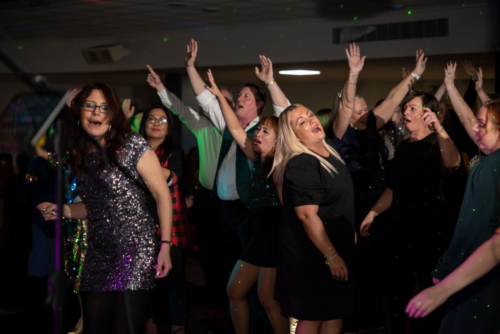 Large group of party guests dancing with arms raised in unison, mid-song on a packed dancefloor with disco lighting.