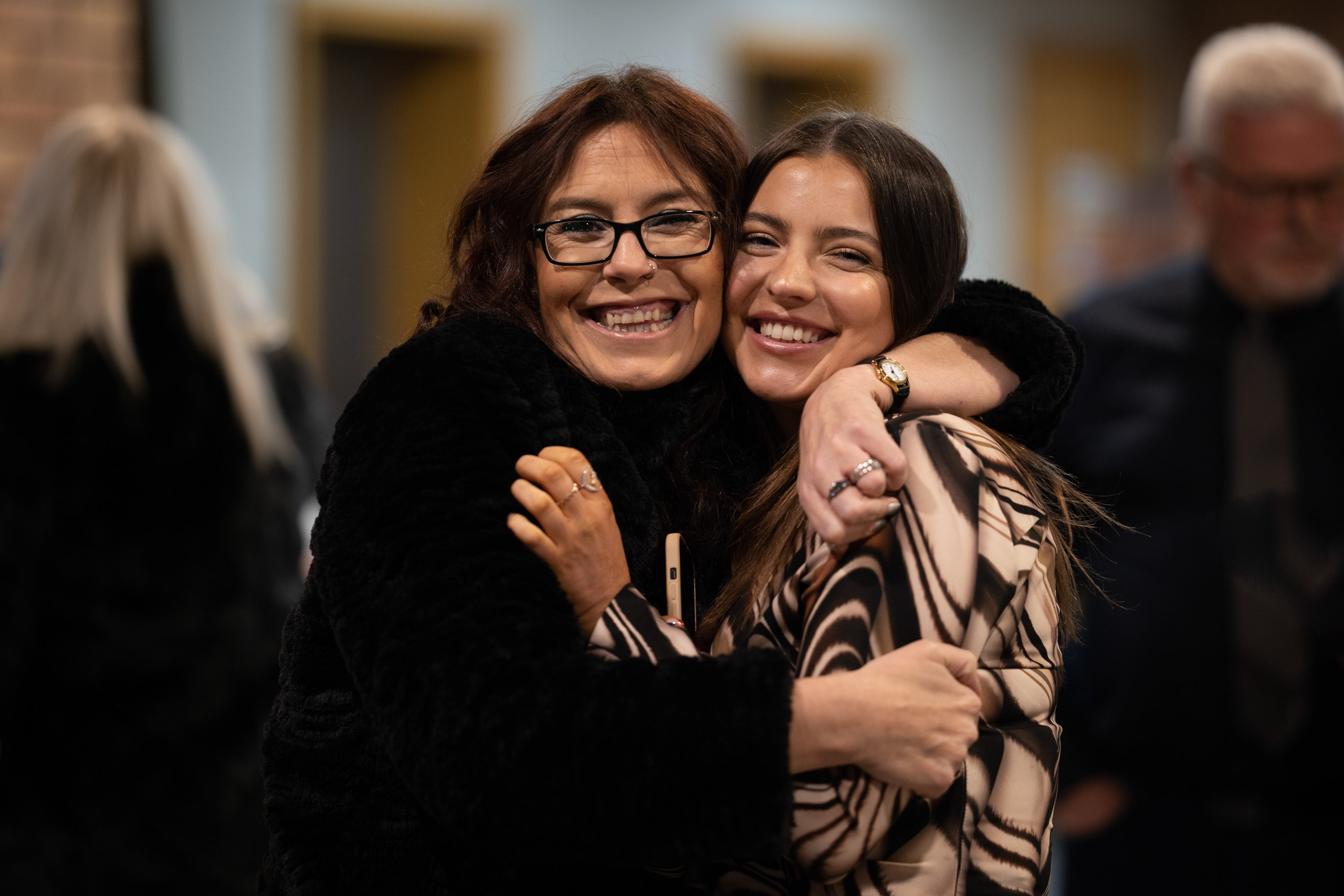 Close-up of two women hugging and smiling at the camera during a celebration, with guests blurred in the background.