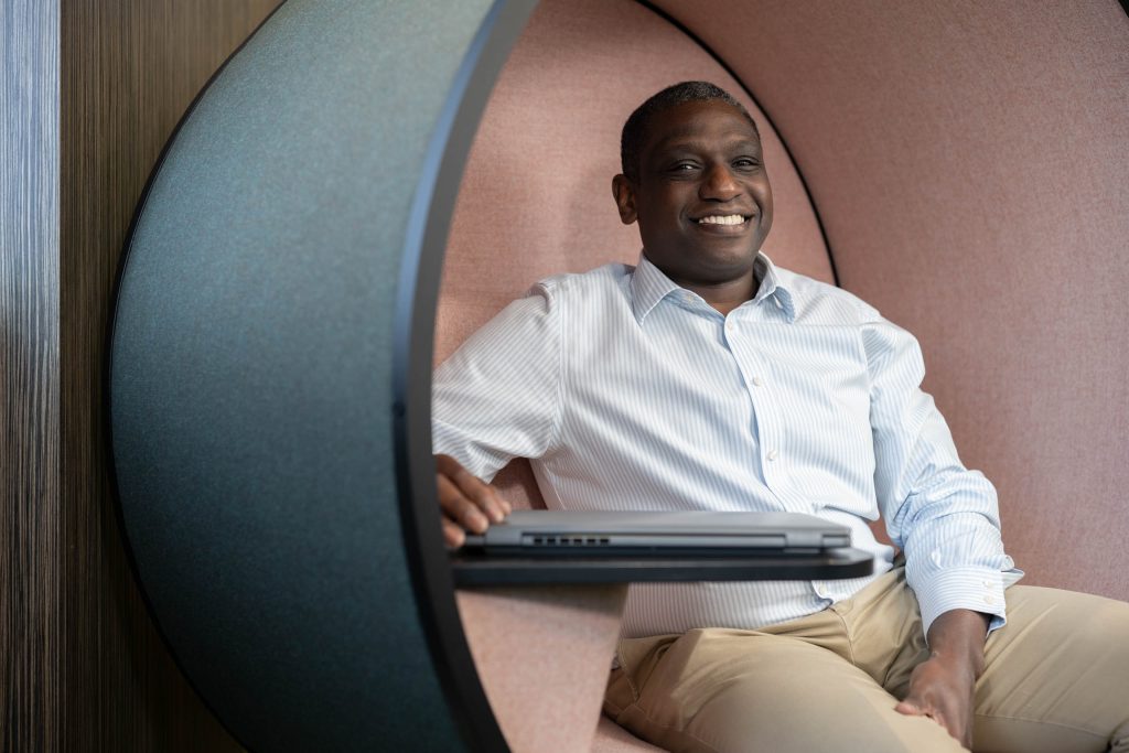 Man in a striped shirt sitting in a pod chair smiling with a laptop in front of him.