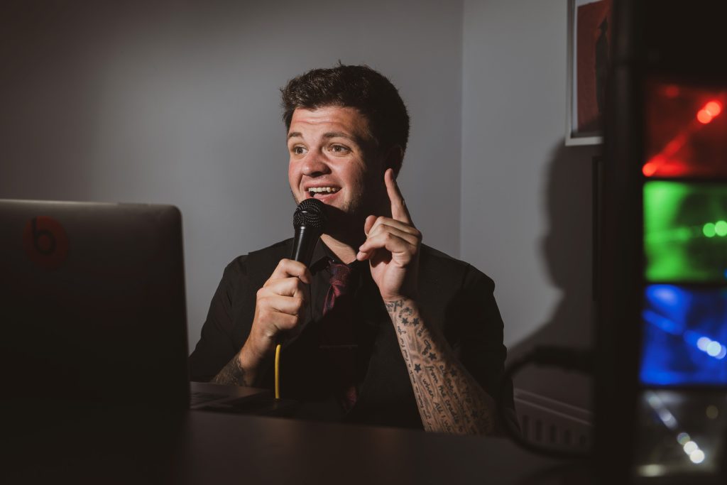 Smiling MC with tattoos speaking into a microphone at an event, framed by colourful lights.