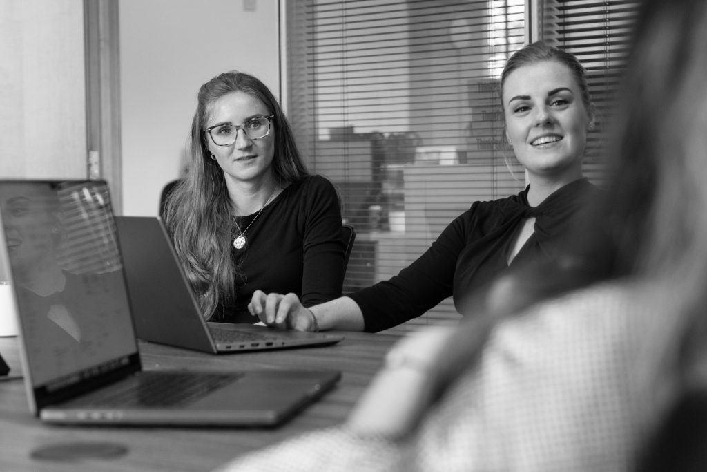 Two women sitting at a desk during a team meeting with laptops in front of them.