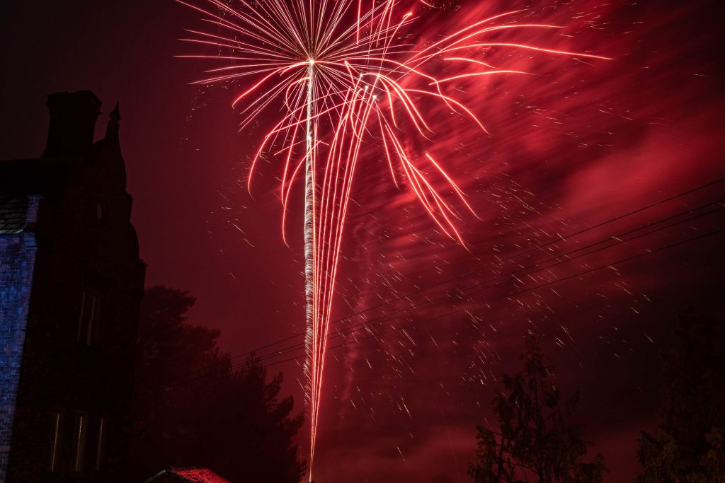 Long-exposure photograph of red fireworks exploding above a silhouetted building and treetops at night.