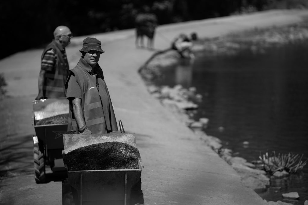 Two volunteers with barrows full of gravel beside a lakeside path, dressed in reflective vests.