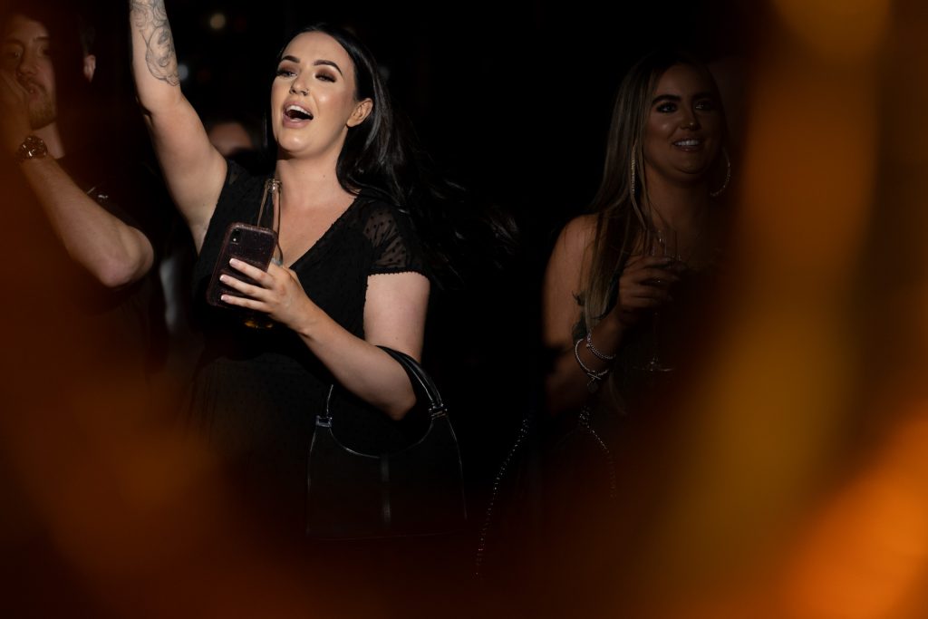 Candid shot of a woman in a black dress smiling with a drink in hand, mid-cheer at a party, surrounded by warm evening light.
