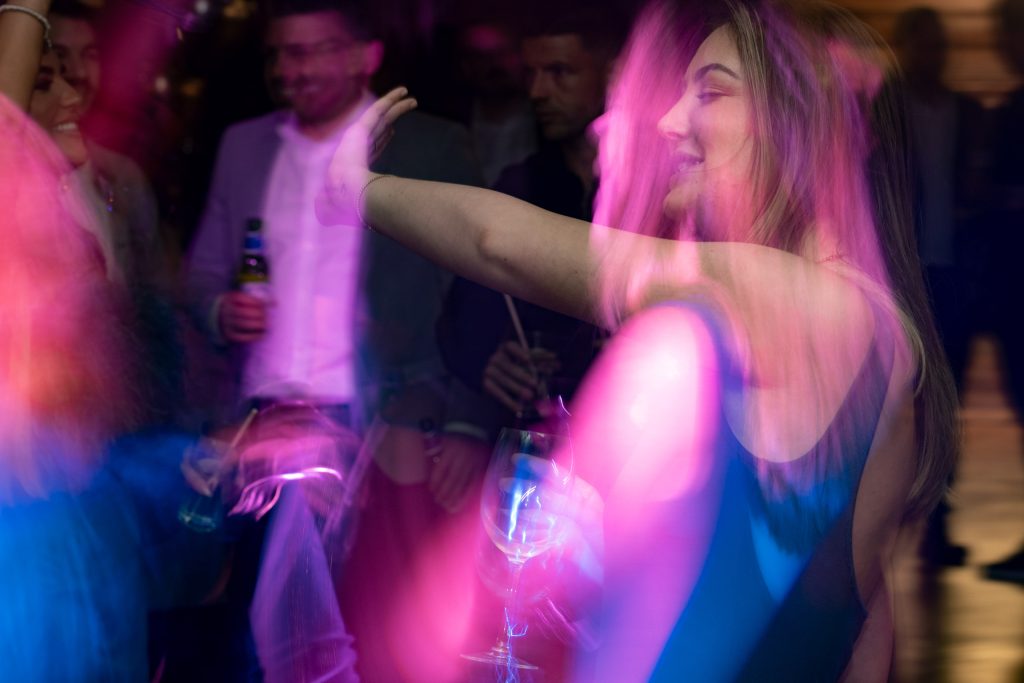 Vibrant long exposure of a woman dancing in pink and purple lights with a glass in hand, surrounded by partygoers.