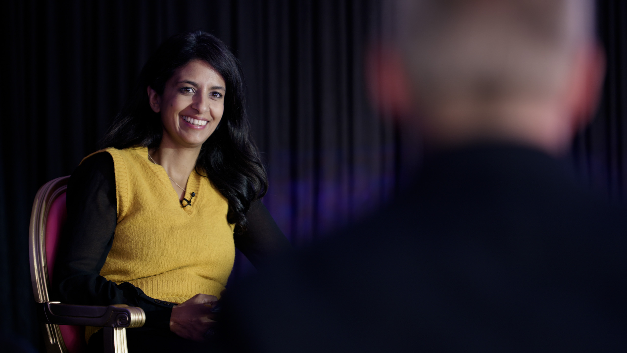 Woman in a yellow top smiling while seated in an interview setting, viewed from behind another person.