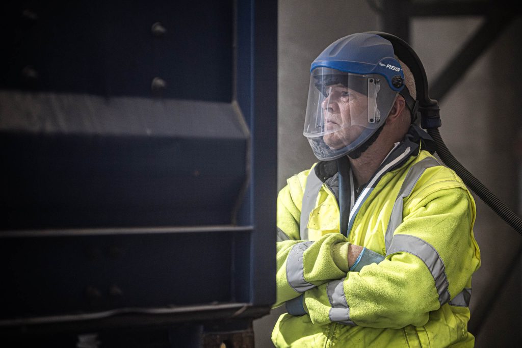 Focused industrial worker in a blue helmet and yellow high-vis jacket, arms crossed amid dusty conditions.
