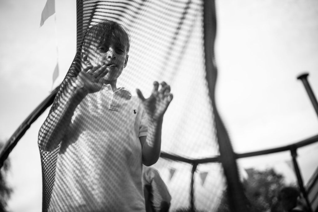 Black and white image of a young boy pressing his hands against the mesh of a trampoline, with sunlight casting patterns on his shirt.