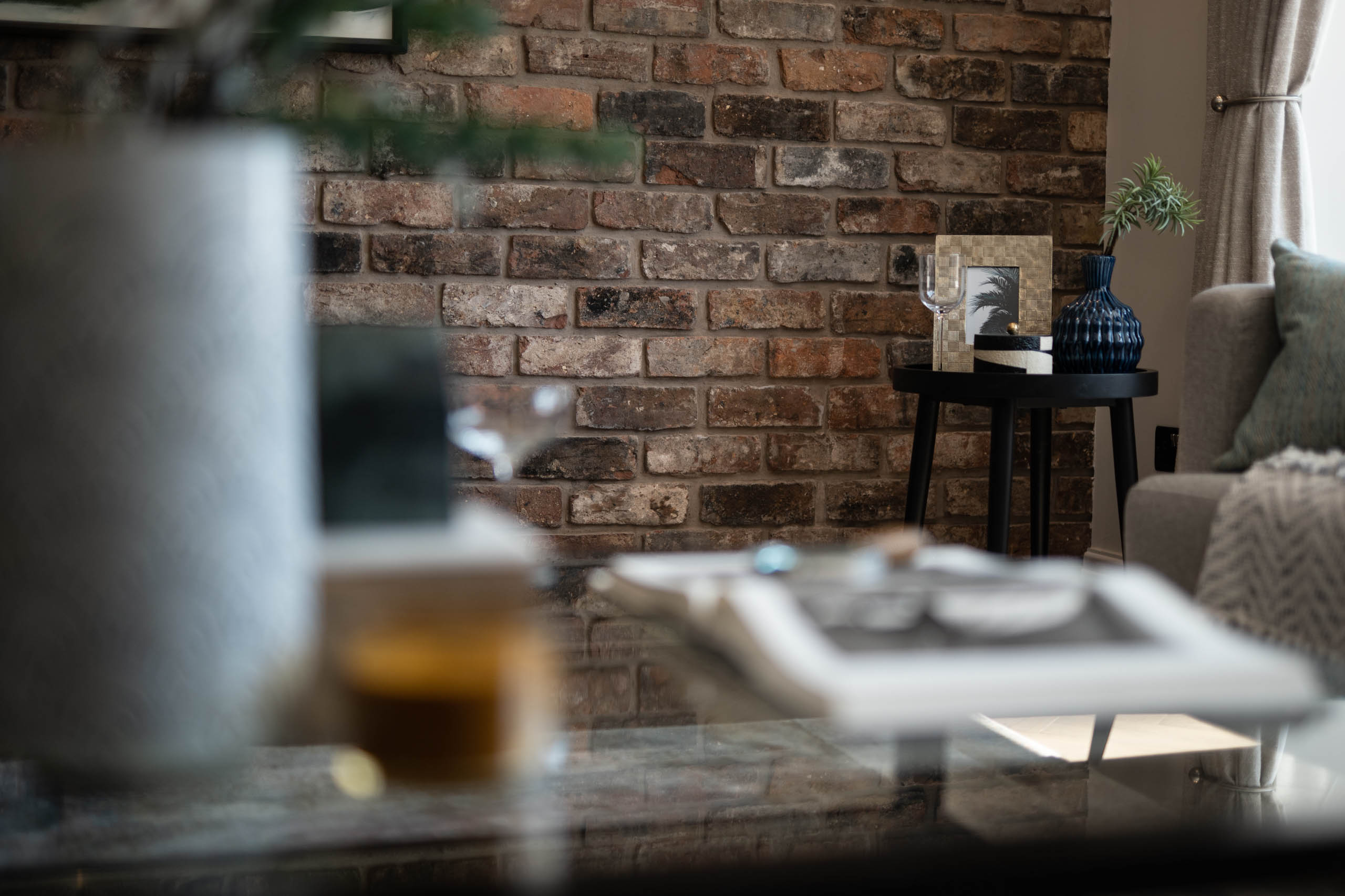 Artistic depth-of-field image showing a brick wall and styled table with glassware and vases in a softly lit room.
