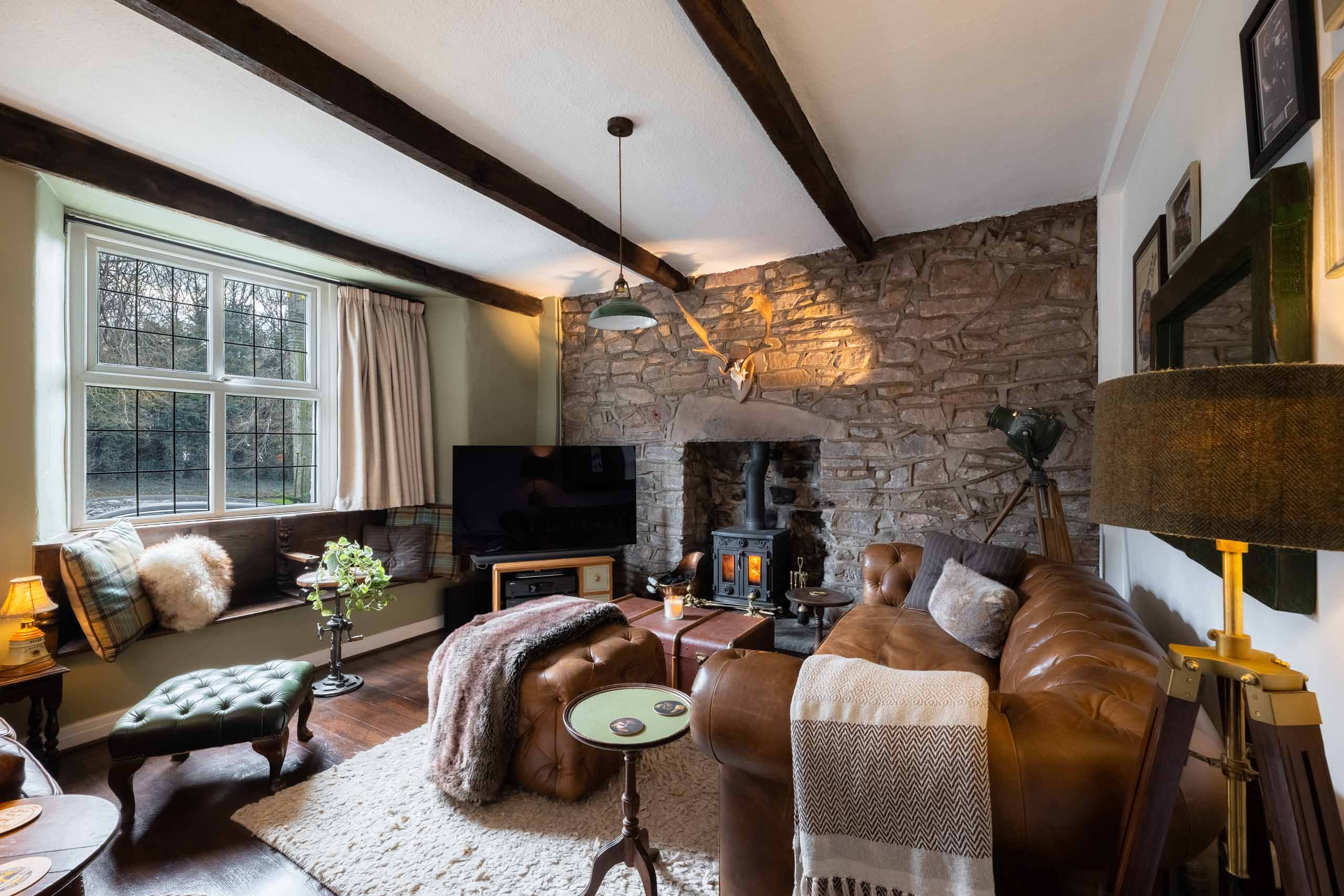 Cosy living room with leather sofa, fur throws, exposed beams, and a lit wood-burning stove against a stone wall.