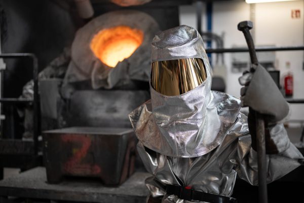 Colour photo of a foundry worker wearing a reflective silver heatproof suit with a gold visor, standing in front of a blazing furnace.