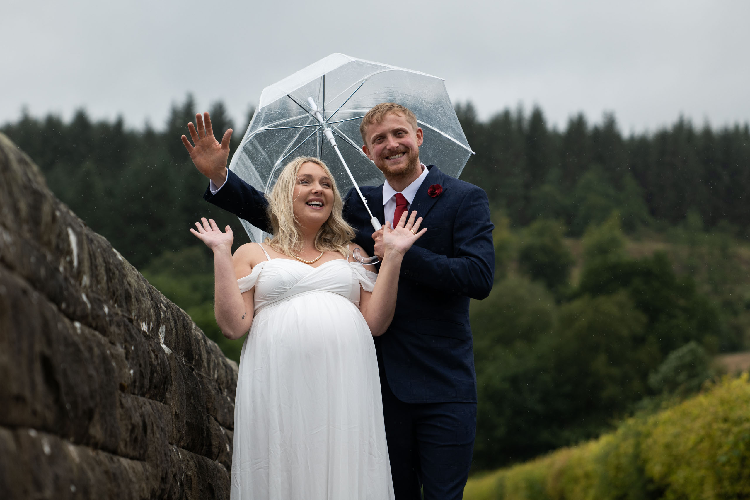 Bride and groom standing under a transparent umbrella on a stone bridge, laughing in the rain.