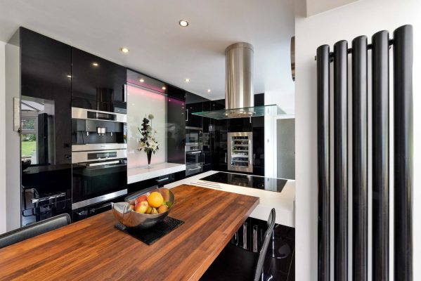 Modern kitchen featuring high-gloss black cabinetry, built-in ovens, wooden breakfast bar, and feature wall lighting.