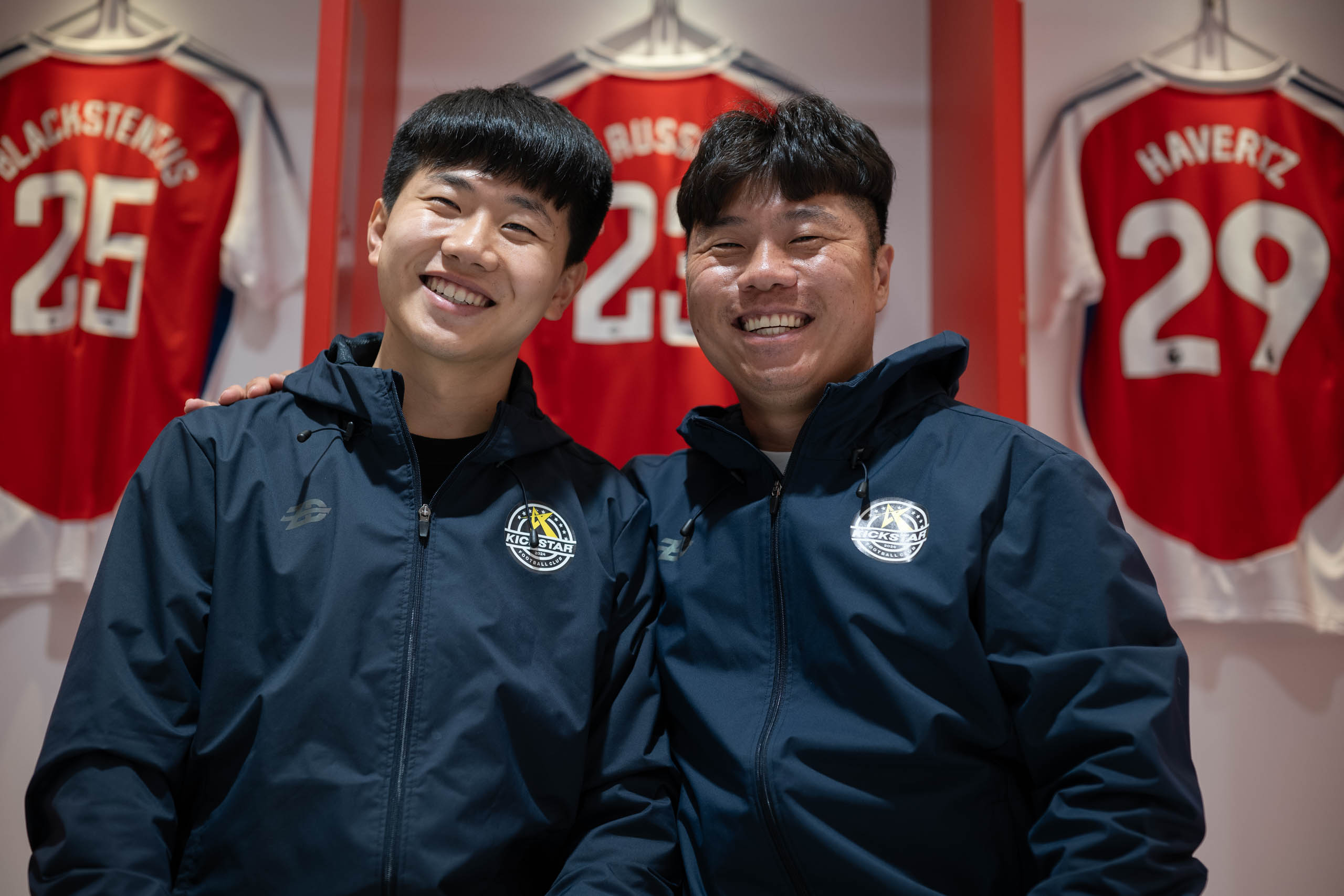 Two football coaches in matching navy jackets posing together in front of Arsenal shirts inside the Emirates locker room.