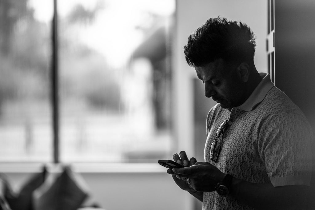 Black and white candid photo of a man leaning against a wall, absorbed in his phone, with soft window light.