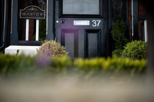 Close-up of a traditional black front door with white trim, showing a 'Moorview' plaque and house number 37 partially obscured by garden plants.