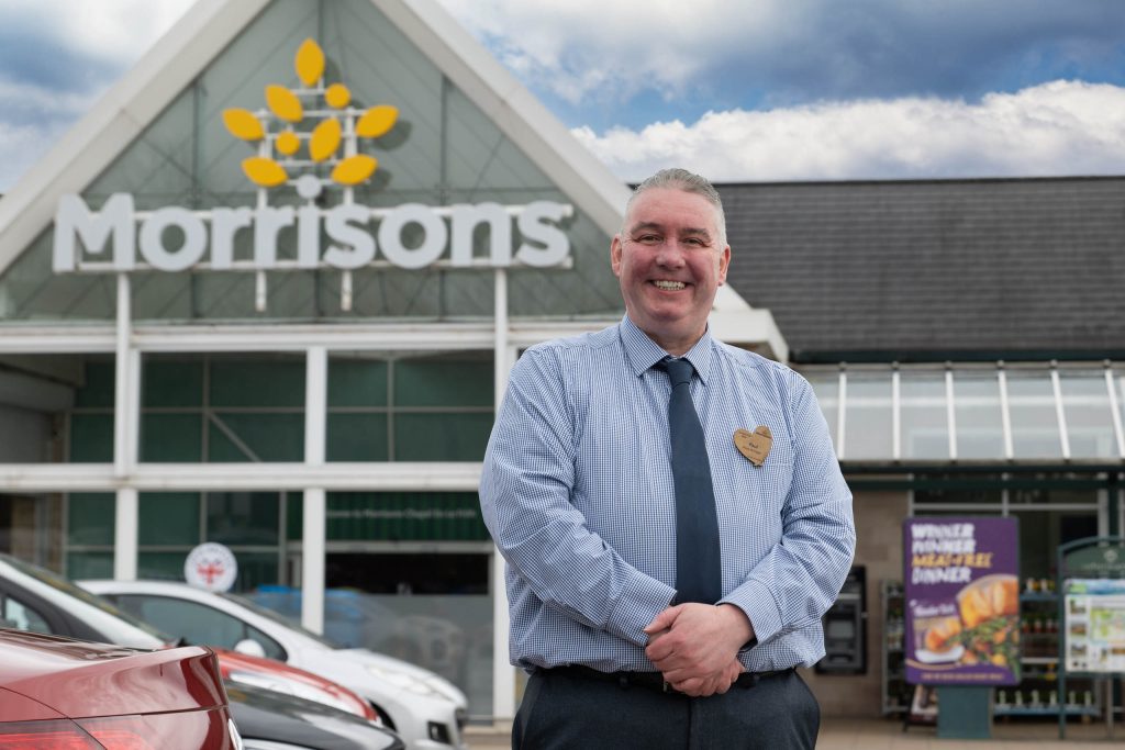 Outdoor portrait of a smiling supermarket manager in uniform standing in front of a Morrisons building with branded signage.
