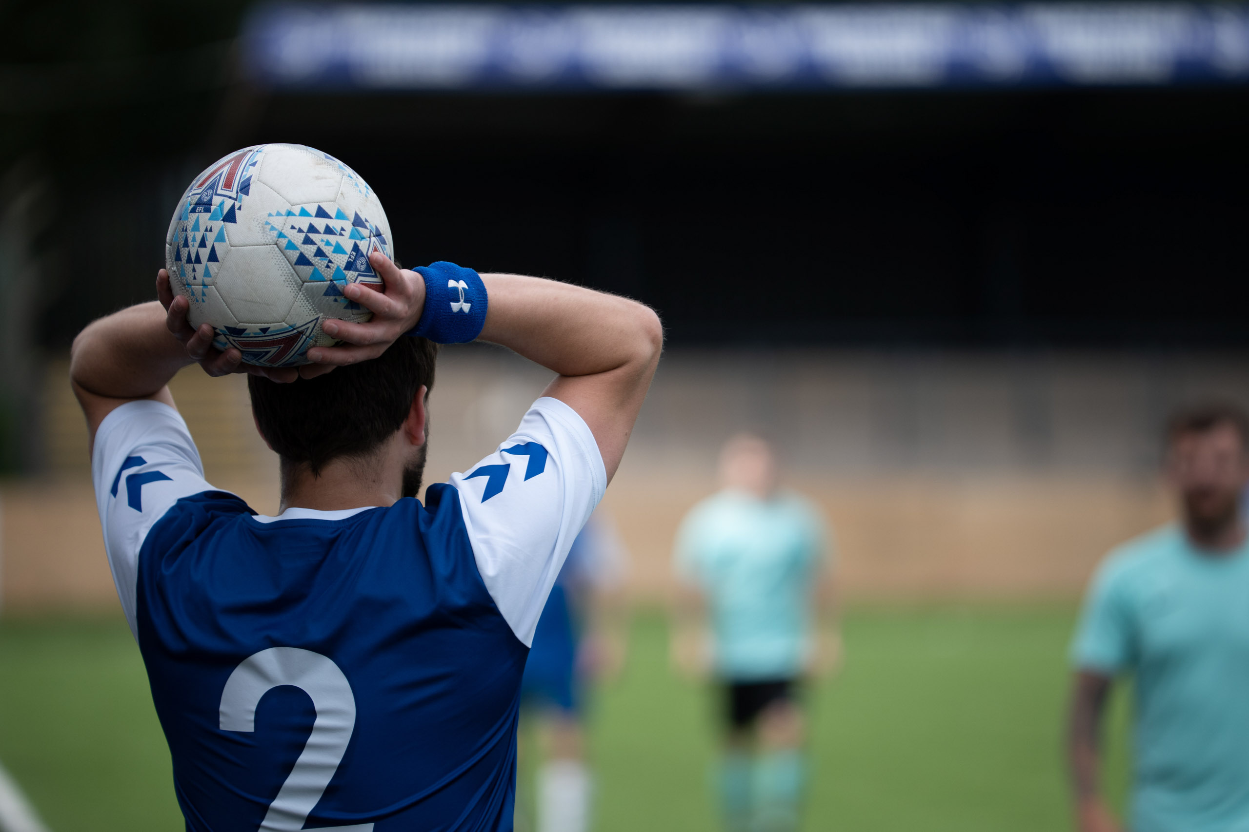 Male footballer with number 2 shirt preparing a throw-in, with teammates and opponents blurred in the background.
