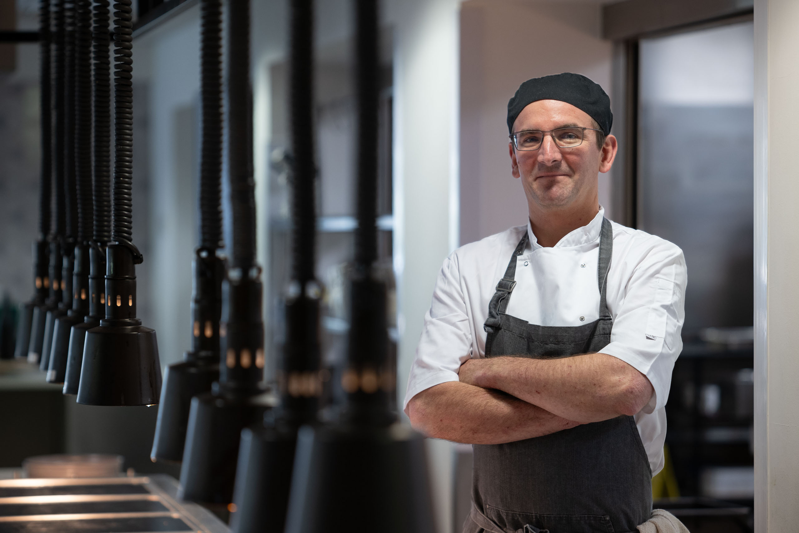 Male chef in a white shirt and apron standing confidently with arms folded between a row of industrial kitchen heat lamps.