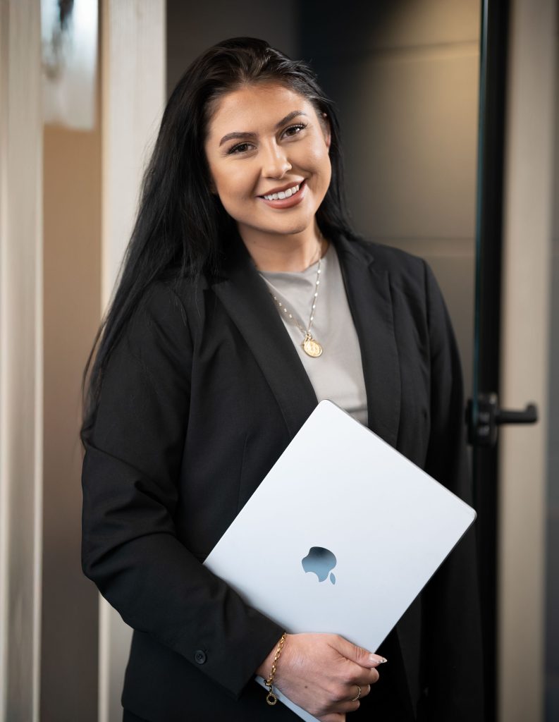Woman in a black blazer smiling while holding a silver MacBook, standing confidently near a glass door.