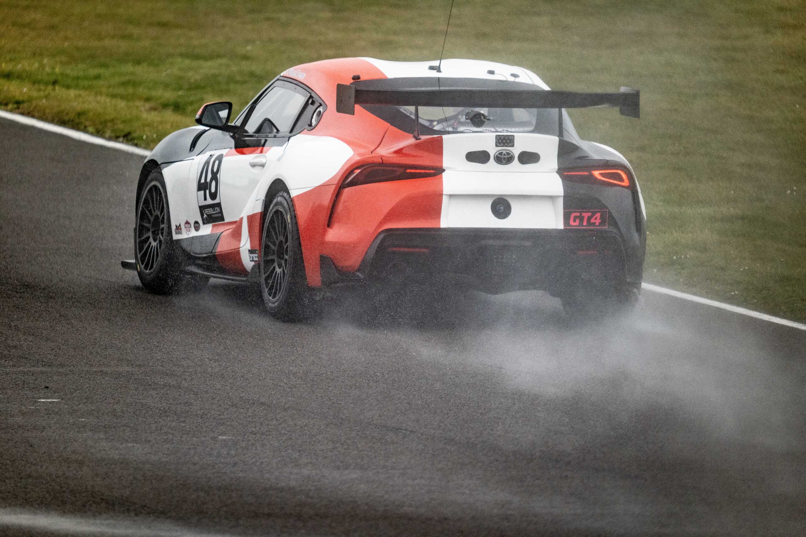 Rear view of a GT4 Toyota Supra racing through a wet track, spraying water into the air behind it.
