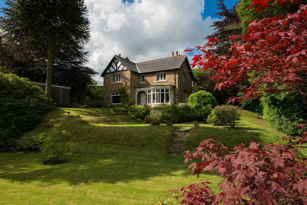 Traditional stone house with gabled roof, bay windows, and mature garden framed by vibrant foliage and a red acer tree.