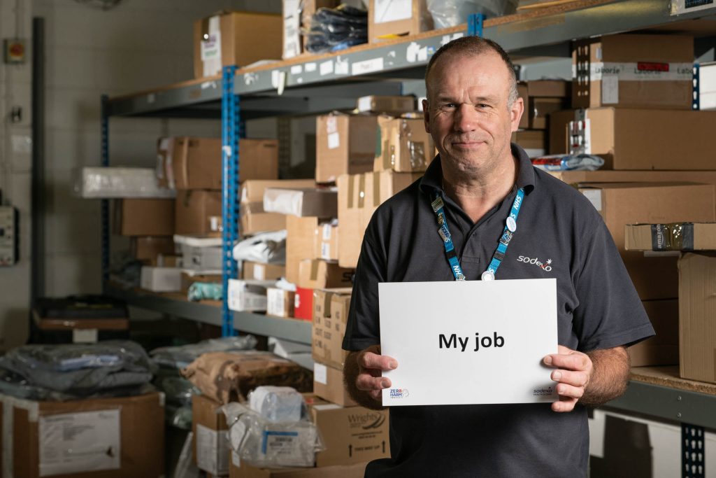 Middle-aged man in a stockroom holding a placard that says ‘My job’, surrounded by boxes and stock shelving.